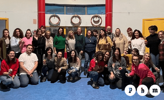 Un grupo numeroso de participantes y formadores del proyecto PBM posa sonriente en el interior de un centro educativo tras una jornada de formación sobre pedagogía Waldorf. Los asistentes, de diversas edades, están distribuidos de pie y agachados, algunos sosteniendo pequeños objetos artesanales. Al fondo se ve una pared blanca con elementos circulares decorativos y el logotipo "PBM" en la esquina inferior derecha.