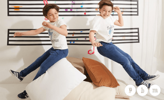 Dos niños saltan con alegría sobre grandes cojines blancos y marrones en una habitación luminosa. Ambos visten camisetas blancas con el lema "Perfectos Imperfectos" y pantalones azules. Al fondo, la pared está decorada con grandes pentagramas musicales y notas de colores, simbolizando la energía de la canción de la Fundación Cadete. En la esquina inferior derecha aparece el logotipo de "PBM".