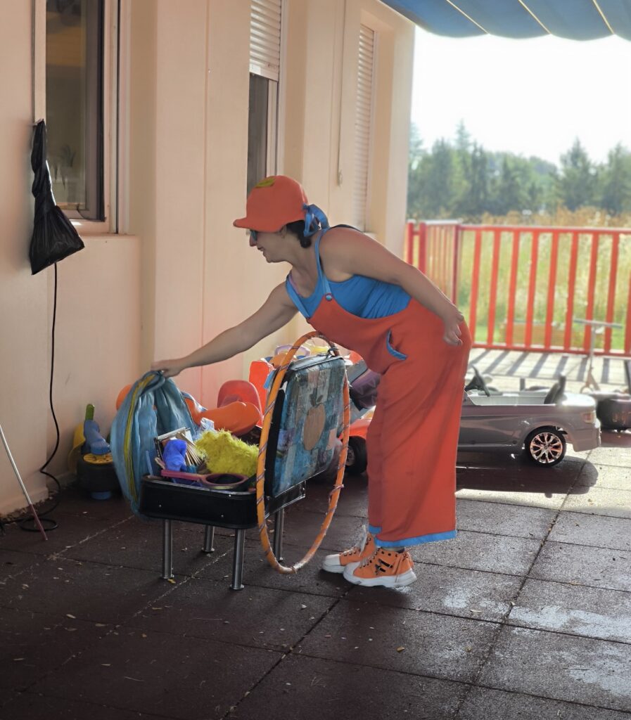 Una animadora infantil vestida con un colorido peto naranja, gorro y gafas azules, realiza un espectáculo de pompas de jabón gigantes frente a un libro de "La Pequeña Oruga Glotona" y juguetes de exterior.