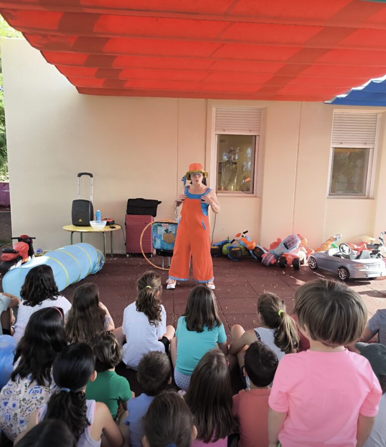 Una animadora infantil vestida con un colorido peto naranja, gorro y gafas azules, realiza un espectáculo de pompas de jabón gigantes frente a un libro de "La Pequeña Oruga Glotona" y juguetes de exterior.