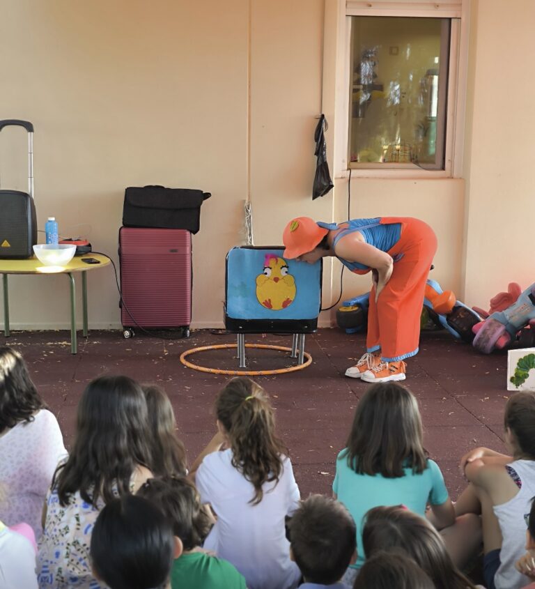 Una animadora infantil vestida con un colorido peto naranja, gorro y gafas azules, realiza un espectáculo de pompas de jabón gigantes frente a un libro de "La Pequeña Oruga Glotona" y juguetes de exterior.
