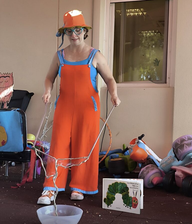 Una animadora infantil vestida con un colorido peto naranja, gorro y gafas azules, realiza un espectáculo de pompas de jabón gigantes frente a un libro de "La Pequeña Oruga Glotona" y juguetes de exterior.