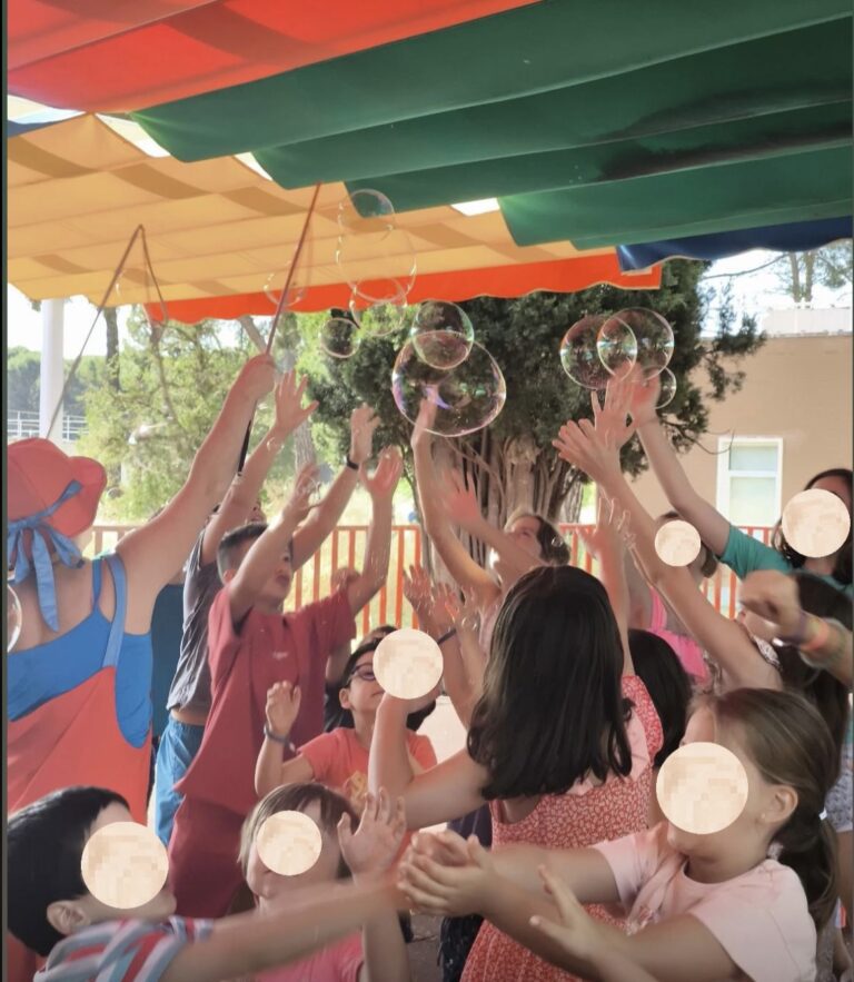 Una animadora infantil vestida con un colorido peto naranja, gorro y gafas azules, realiza un espectáculo de pompas de jabón gigantes frente a un libro de "La Pequeña Oruga Glotona" y juguetes de exterior.