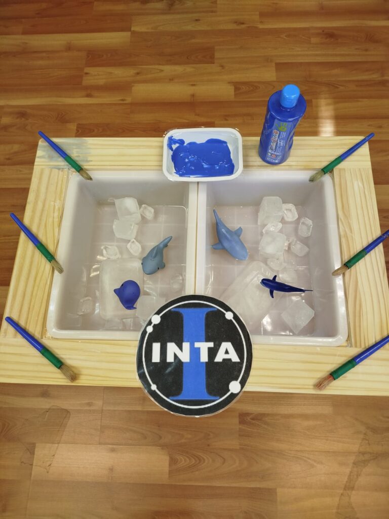 Explorando el cambio de estado del hielo mientras aplican pintura azul con pinceles. En la bandeja se ven juguetes marinos y hielo derritiéndose, creando una experiencia sensorial completa.