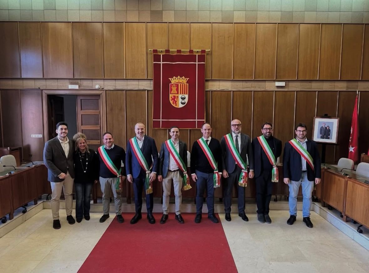Fotografía de grupo en un salón de plenos institucional durante la visita de una delegación de alcaldes de la región de Reggio Emilia, Italia, pertenecientes a Progettinfanzia de Bassa Reggiana. En la imagen aparecen representantes municipales junto a responsables de la escuela, varios de ellos portando bandas oficiales con los colores de la bandera italiana. Dos de los asistentes sostienen placas o diplomas conmemorativos del encuentro. Al fondo se observa el escudo institucional y banderas oficiales, en el marco de un intercambio pedagógico del proyecto Erasmus.