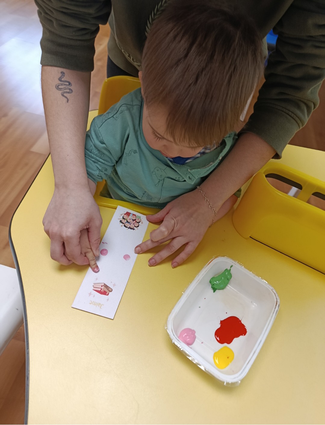 Primer plano de una educadora ayudando a un bebé a poner su huella con pintura rosa en un marcapáginas personalizado.
