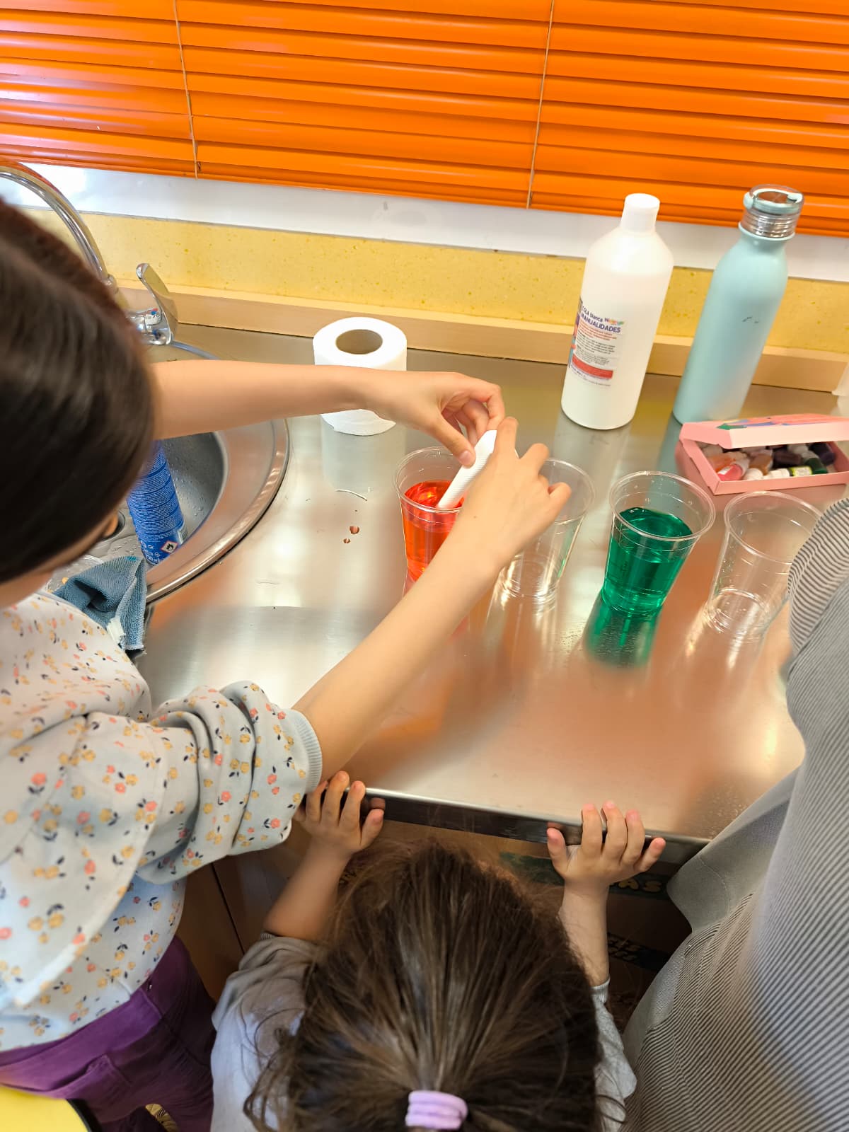 Niños manipulando vasos con agua de colores y papel absorbente en un taller educativo sobre una mesa metálica.