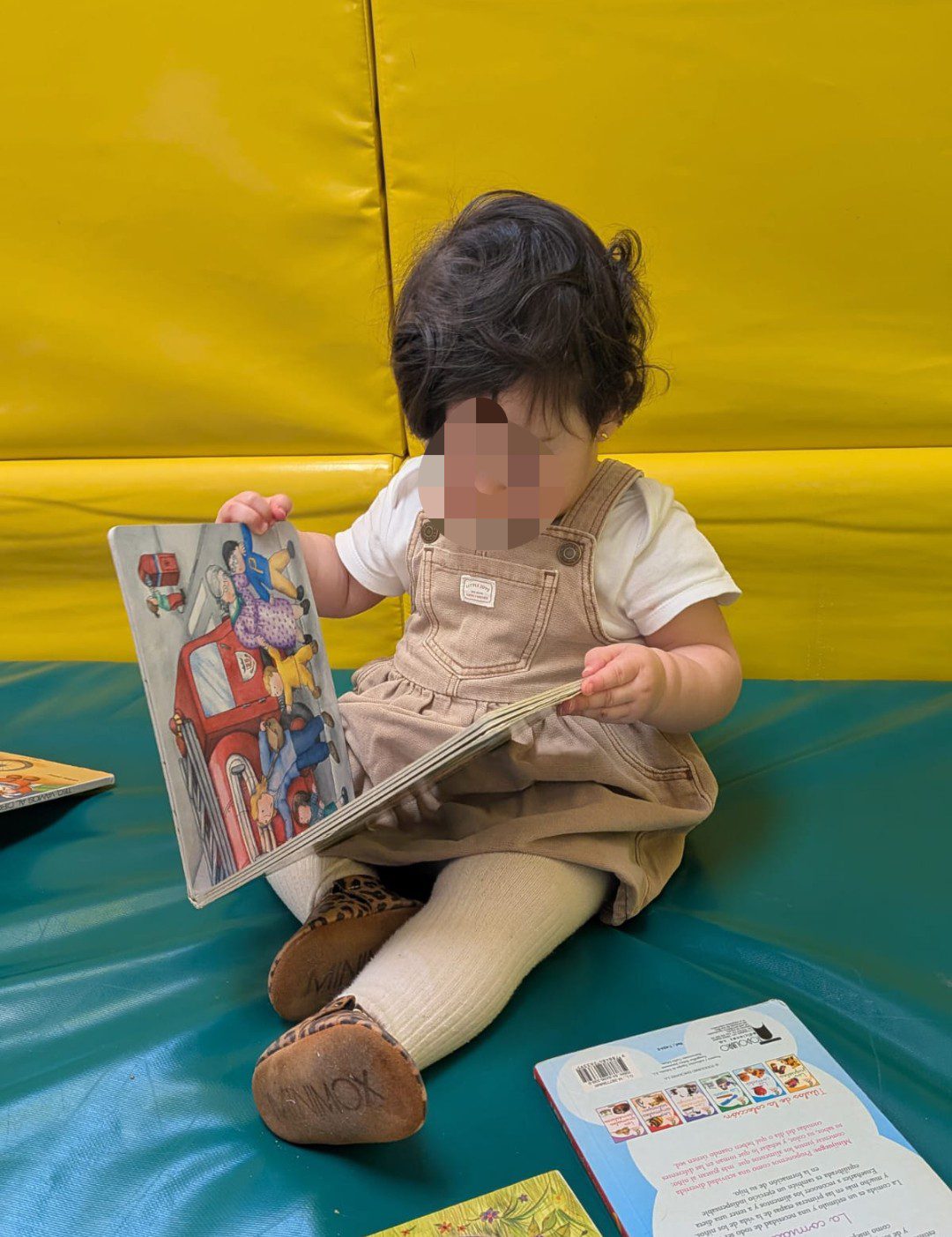 Bebé sentado en una colchoneta verde de la escuela La Casita, vestido con un peto beige, sosteniendo y mirando con atención un libro infantil abierto.