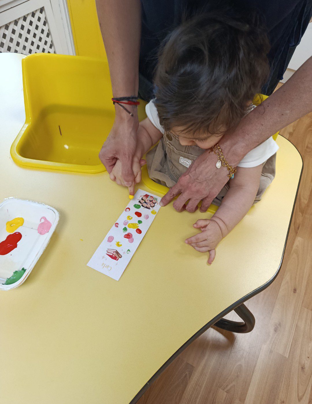 Primer plano de una profesora ayudando a una niña a pintar un marcapáginas personalizado con pintura de dedos en una escuela infantil.
