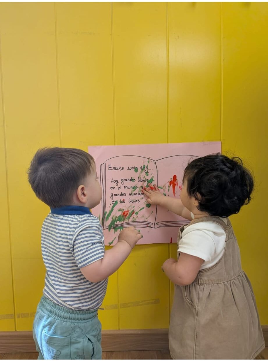 Dos niños pequeños compartiendo y explorando juntos un mural de lectura en el aula.