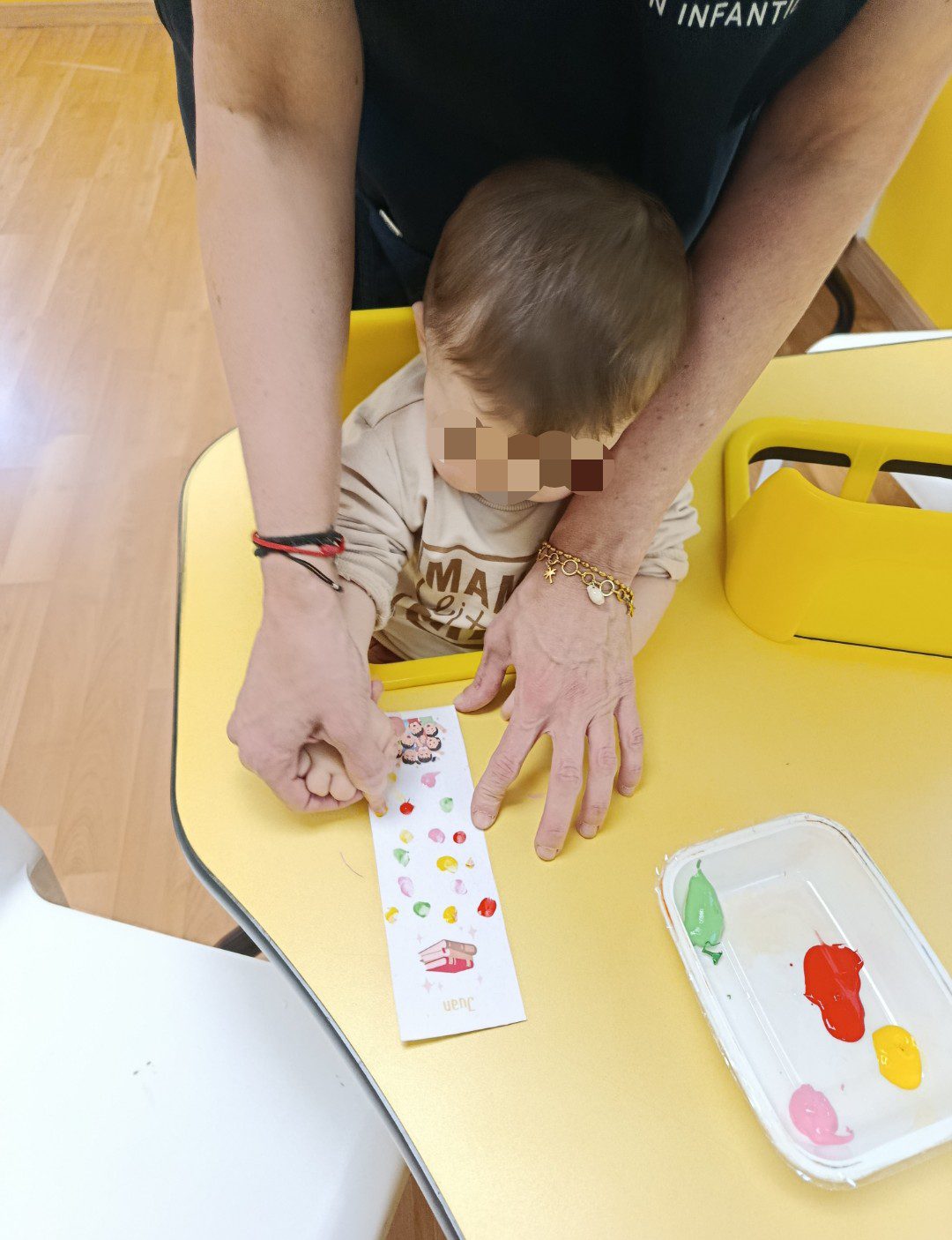 Educadora infantil guiando la mano de un niño pequeño para pintar con los dedos sobre un marcapáginas en un aula de Torrejón de Ardoz.