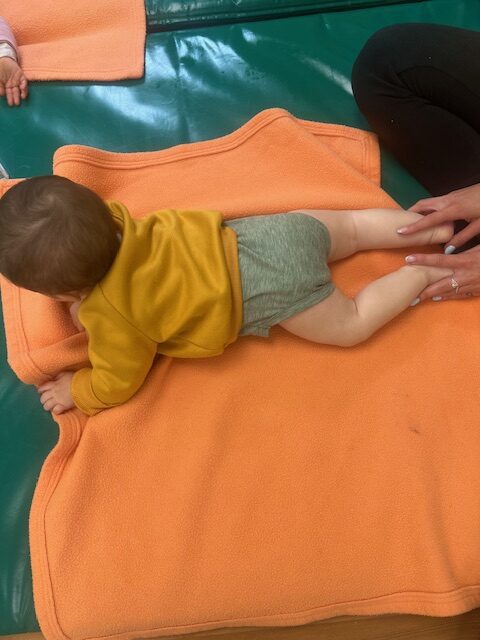 Un bebé de 0 a 1 año tumbado boca abajo sobre una manta naranja mientras una educadora le da un masaje relajante en las piernas y pies en la Escuela Infantil La Casita.