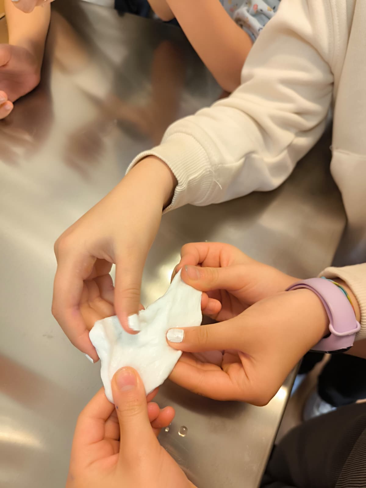 Detalle de manos infantiles manipulando y estirando una masa blanca viscosa sobre una superficie metálica en un taller sensorial.