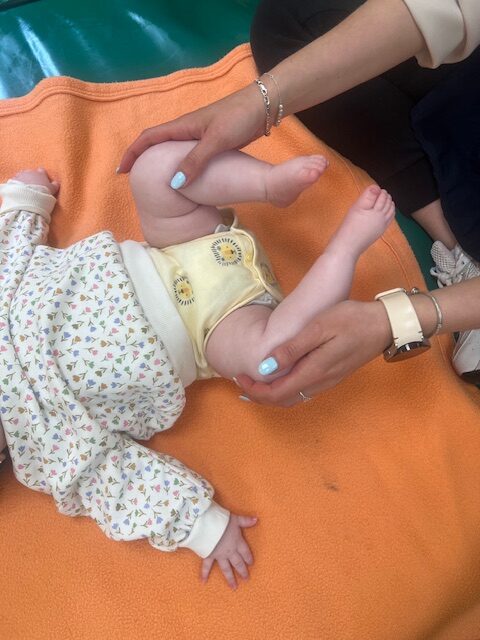 Primer plano de una educadora infantil realizando ejercicios de flexión y masaje en las piernas de un bebé sobre una manta naranja en la Escuela Infantil La Casita.