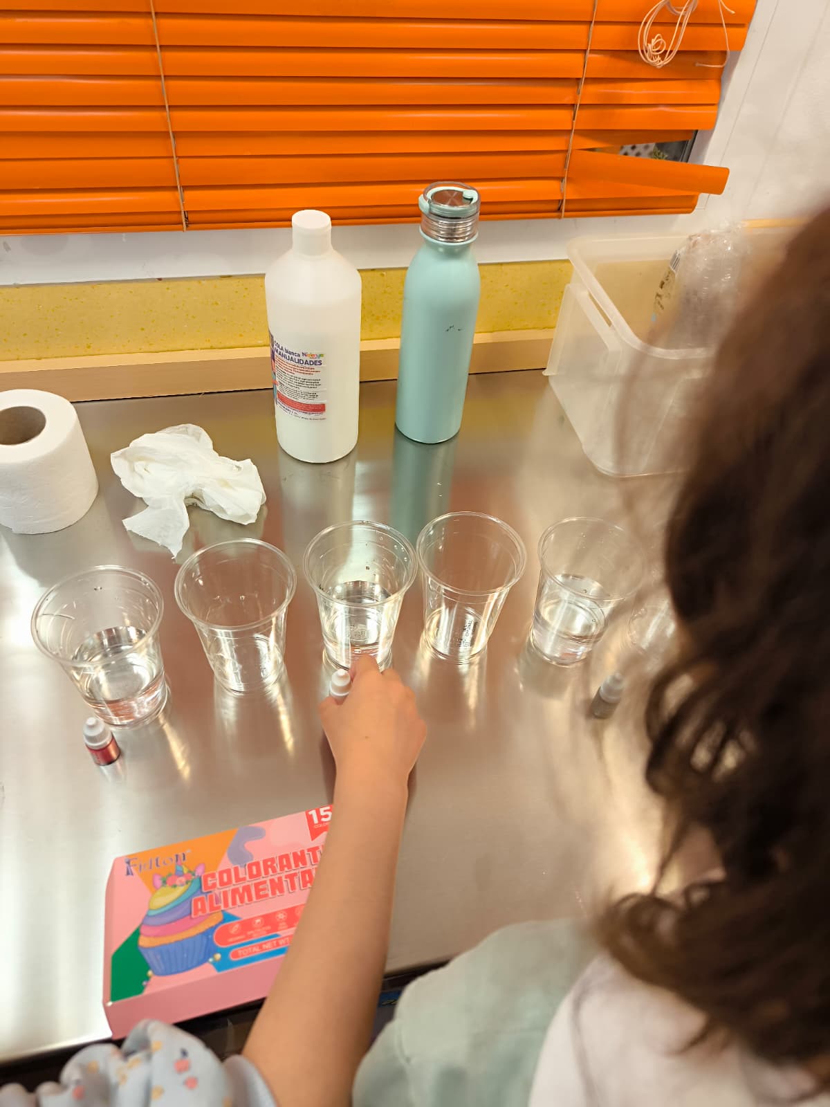 Niña preparando vasos de agua con colorante alimentario en una mesa de metal para un experimento de ciencia infantil.