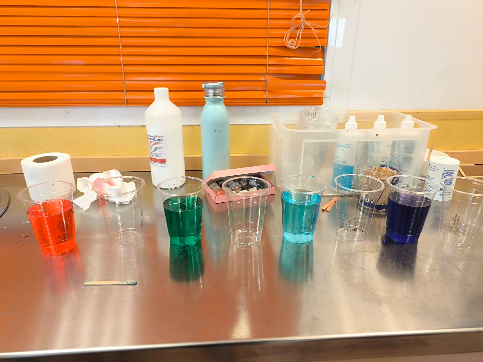 Fila de vasos con agua de colores vibrantes preparados para un experimento de estimulación visual en un aula escolar.