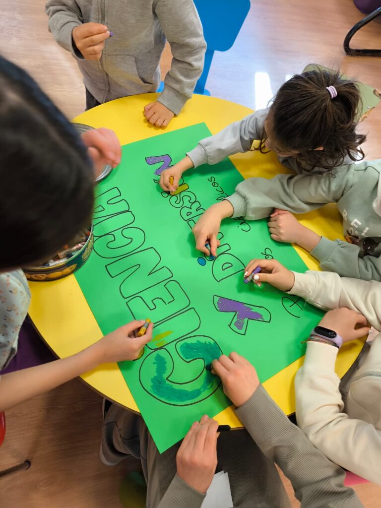 Grupo de niños coloreando juntos un cartel verde sobre una mesa amarilla en un aula de educación infantil.