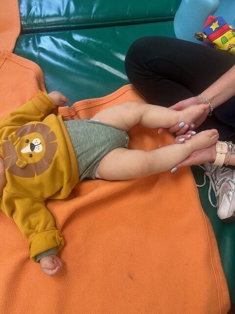 Un bebé de 0 a 1 año, vestido con una sudadera de león y tumbado boca arriba sobre una manta naranja, recibe un suave masaje en los pies de una educadora en la Escuela Infantil La Casita.