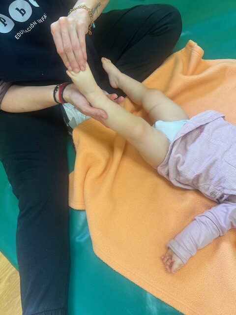 Primer plano de las manos de una educadora de la Escuela Infantil La Casita realizando un masaje suave y rítmico en los pies y piernas de un bebé, tumbado sobre una manta naranja durante un taller de bienestar.