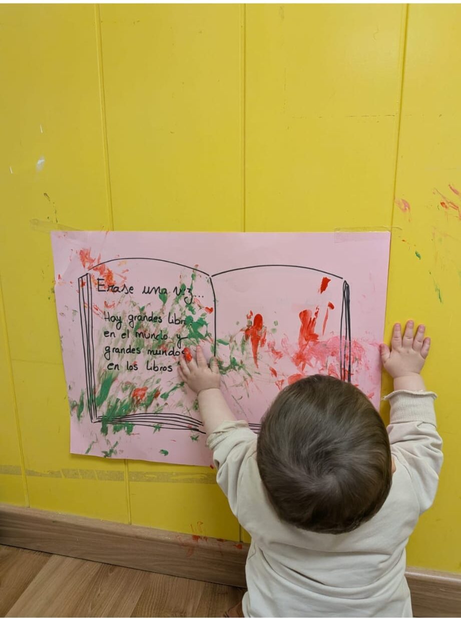 Bebé participando en un mural colectivo con pintura de dedos en la pared de la escuela infantil.
