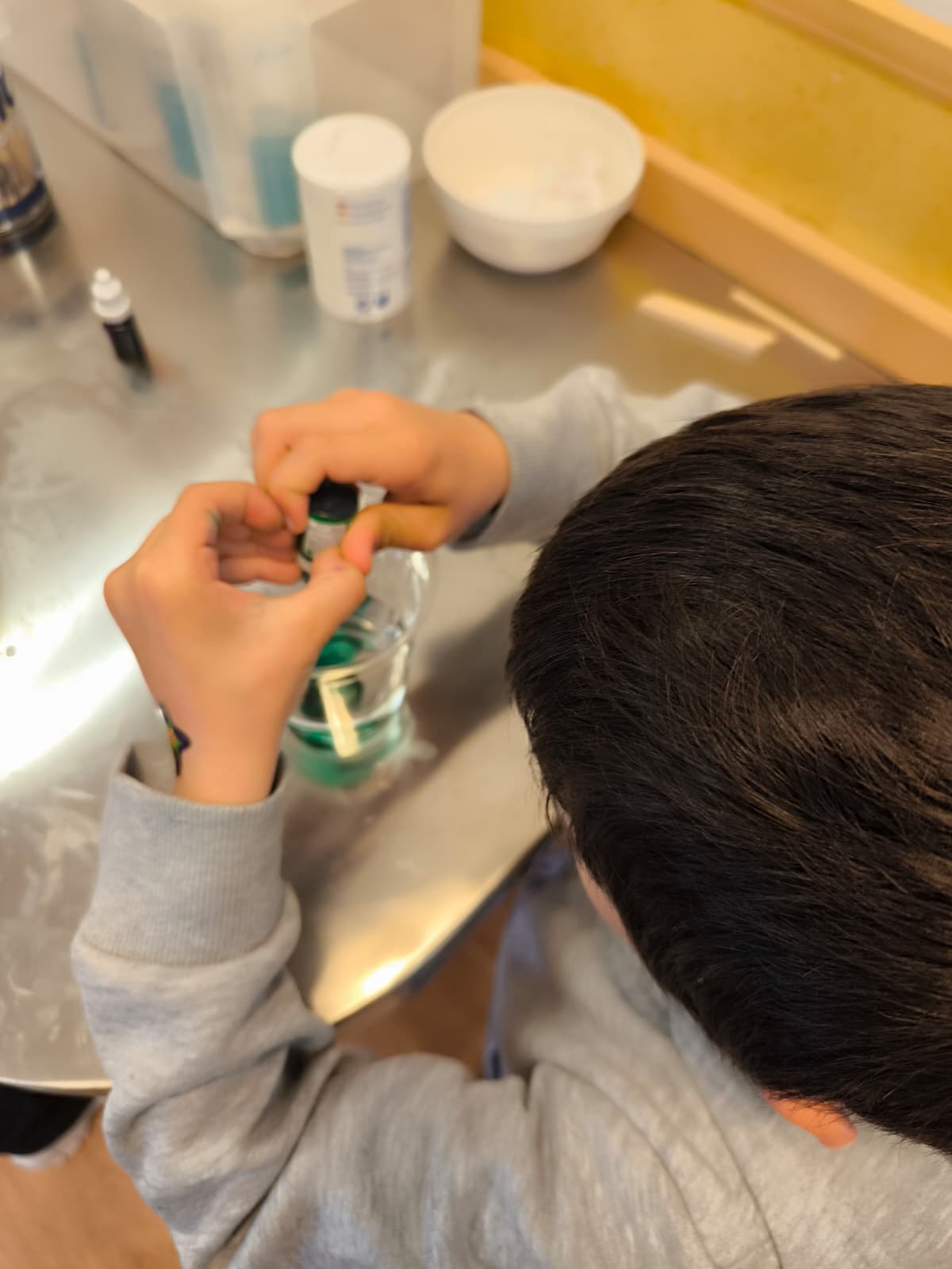 Un niño pequeño concentrado añadiendo gotas de colorante verde a un vaso de agua sobre una mesa de metal.