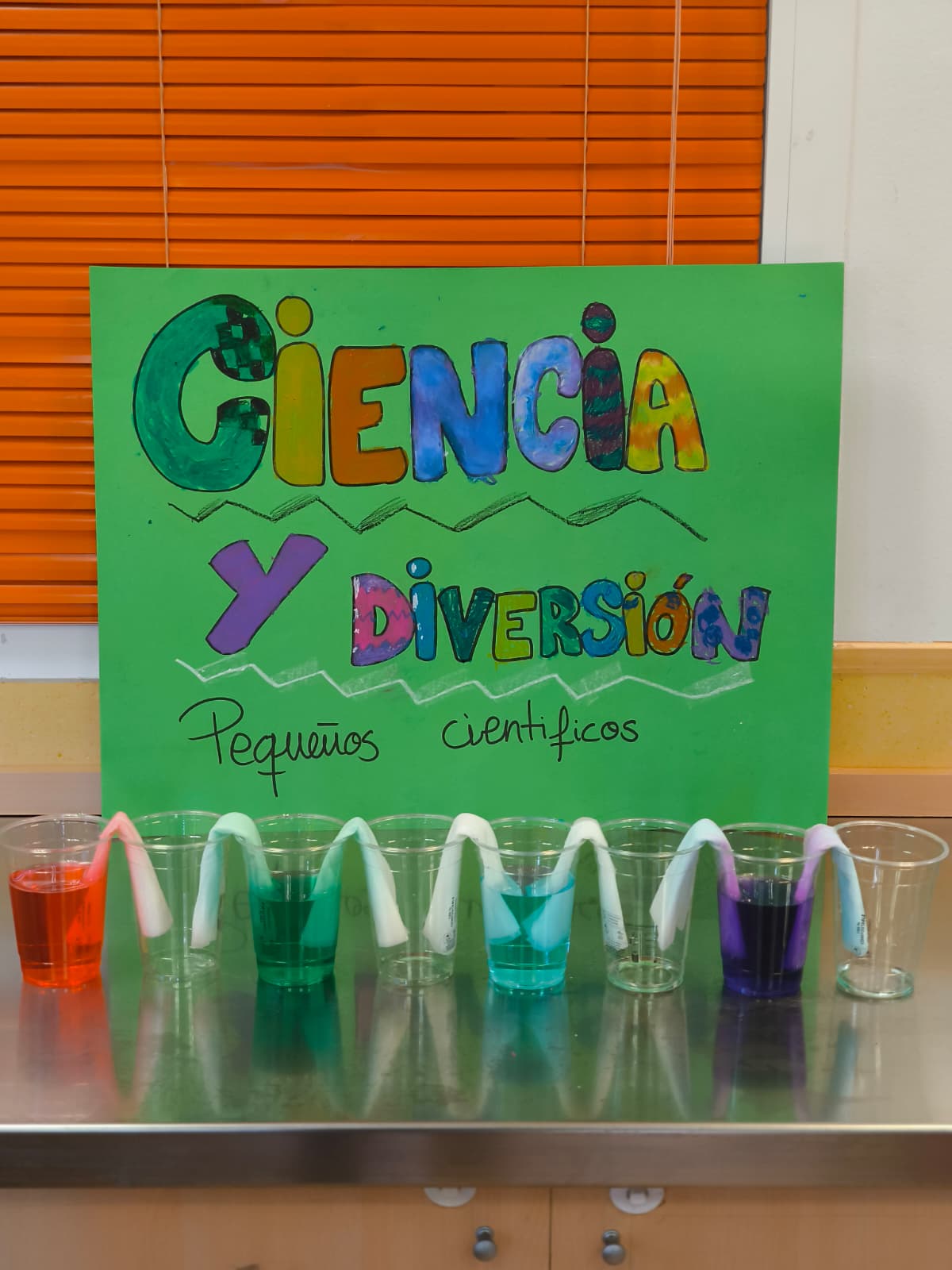 Experimento escolar de vasos con agua de colores y servilletas de papel para demostrar la capilaridad frente a un cartel de "Ciencia y Diversión".