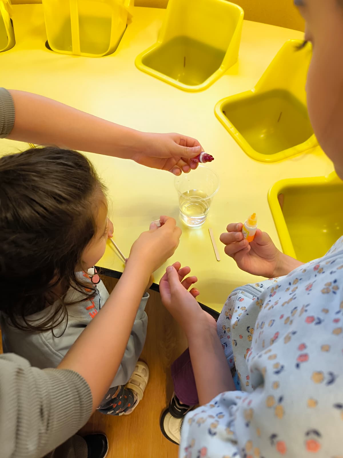 Niños colaborando en una mesa amarilla para mezclar colorantes en vasos de agua durante un taller escolar.